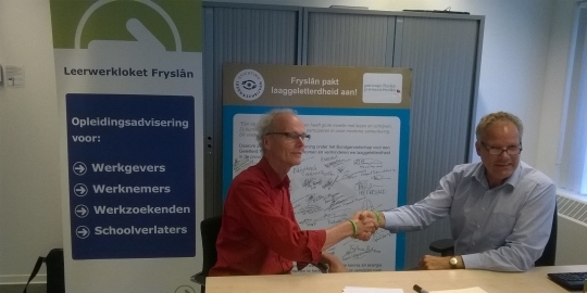 Jitze Bok schudt hand met de vertegenwoordiger van Stichting lezen en schrijven na het ondertekenen van het bondgenootschap. Op de banner van Leerwerkloket Friesland staat: Leerwerkloket Fryslan. Opleidingsadvisering voor werkgevers, werknemers, werkzoekenden en schoolverlaters.