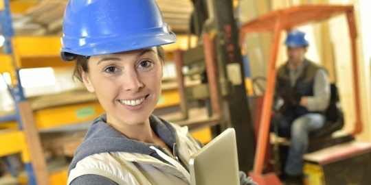 vrouw met bouwhelm, achter haar zit een mannelijke collega in een werkmachine